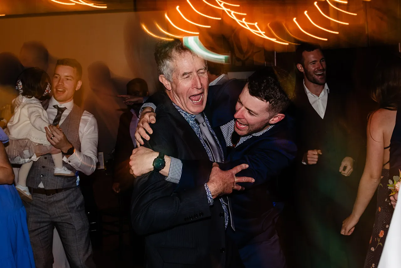 Two men in suits hugging and laughing at a lively indoor event with blurred lights overhead.