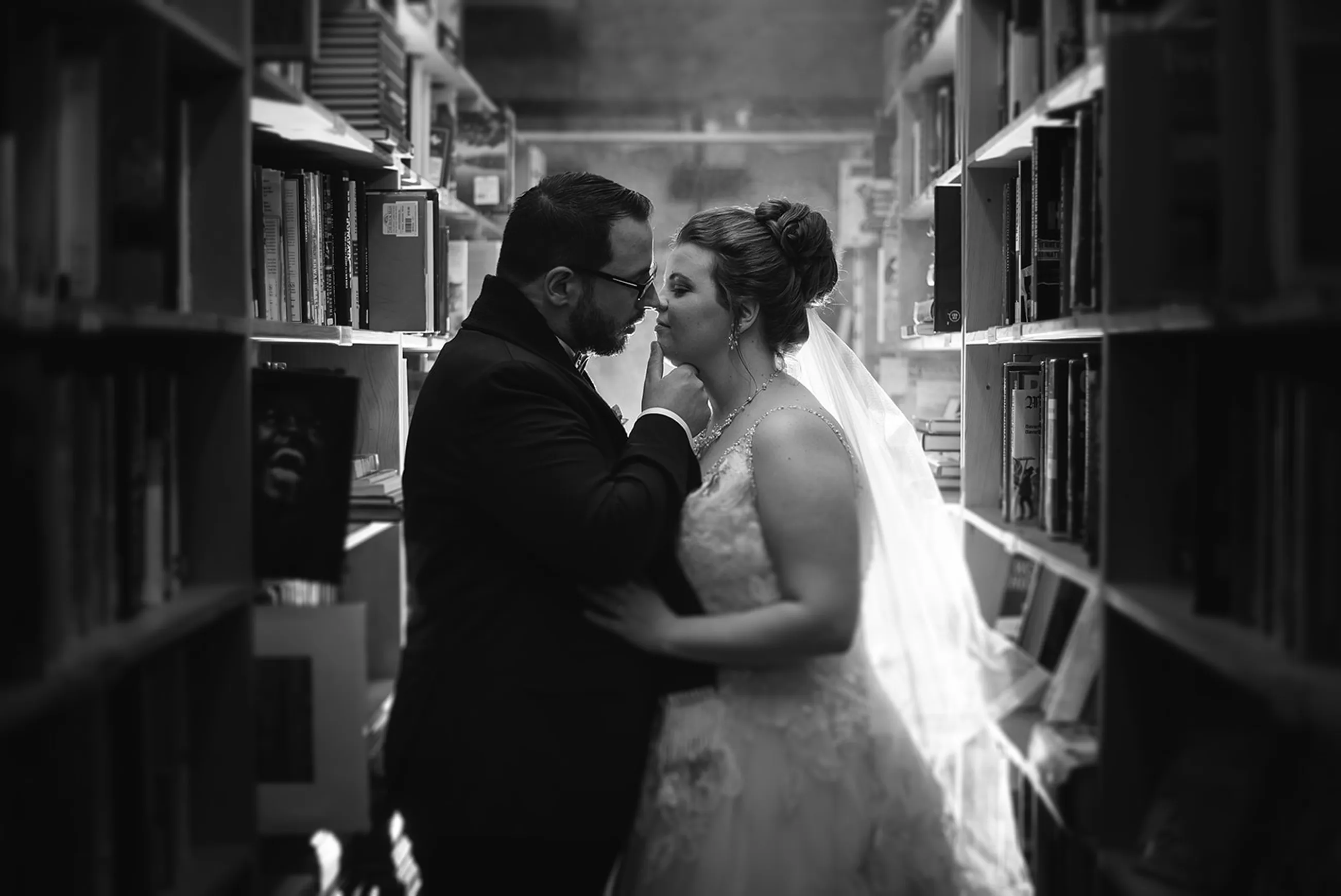Bride and groom in wedding attire sharing an intimate moment between library bookshelves in black and white.