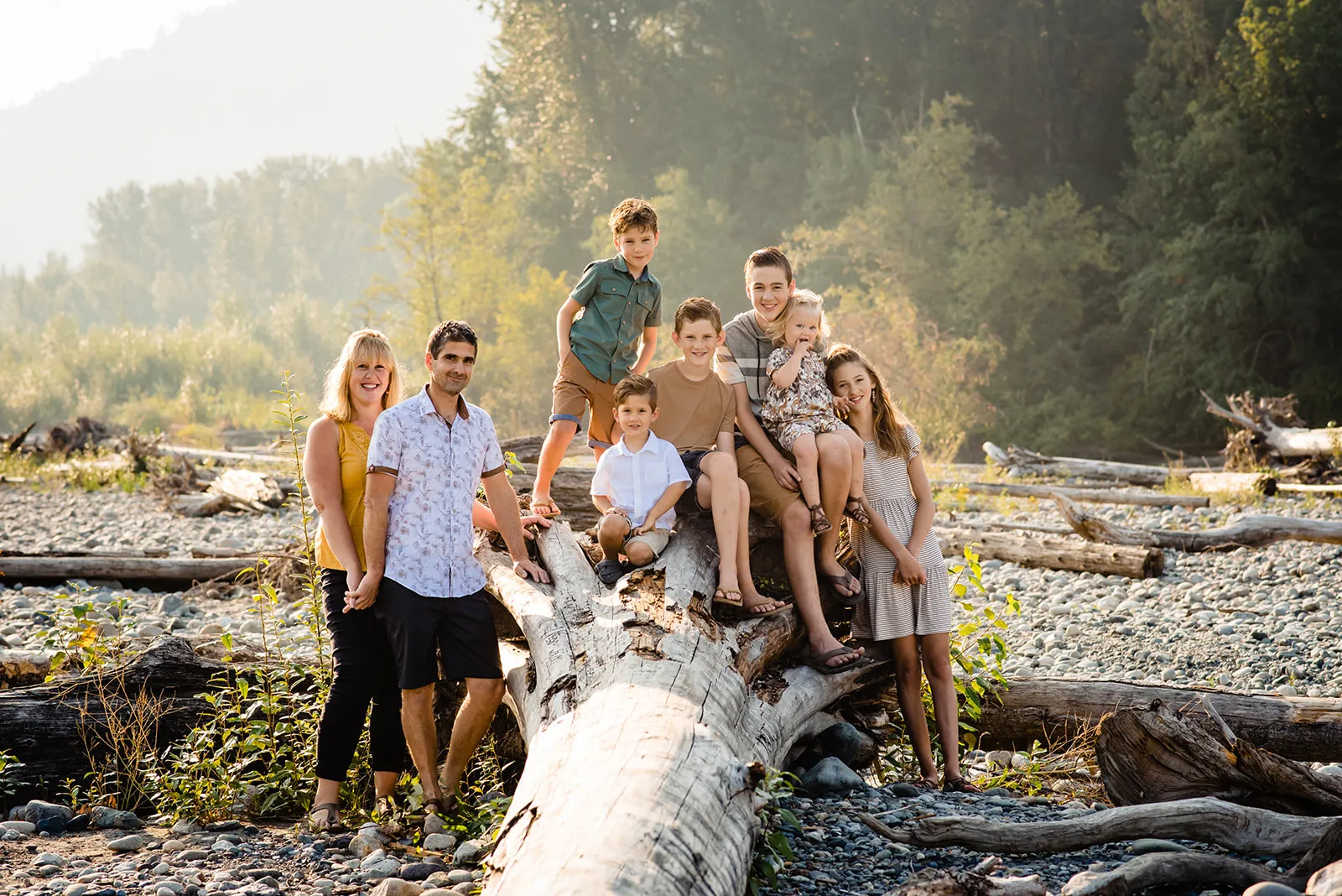 Family of eight posing on and around a large fallen tree in a sunny forest clearing with rocks.