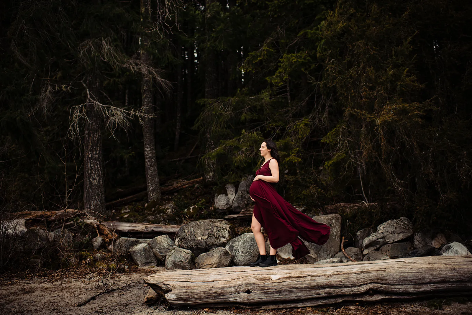 Pregnant woman in a flowing maroon dress walking on a large log in a forest setting.