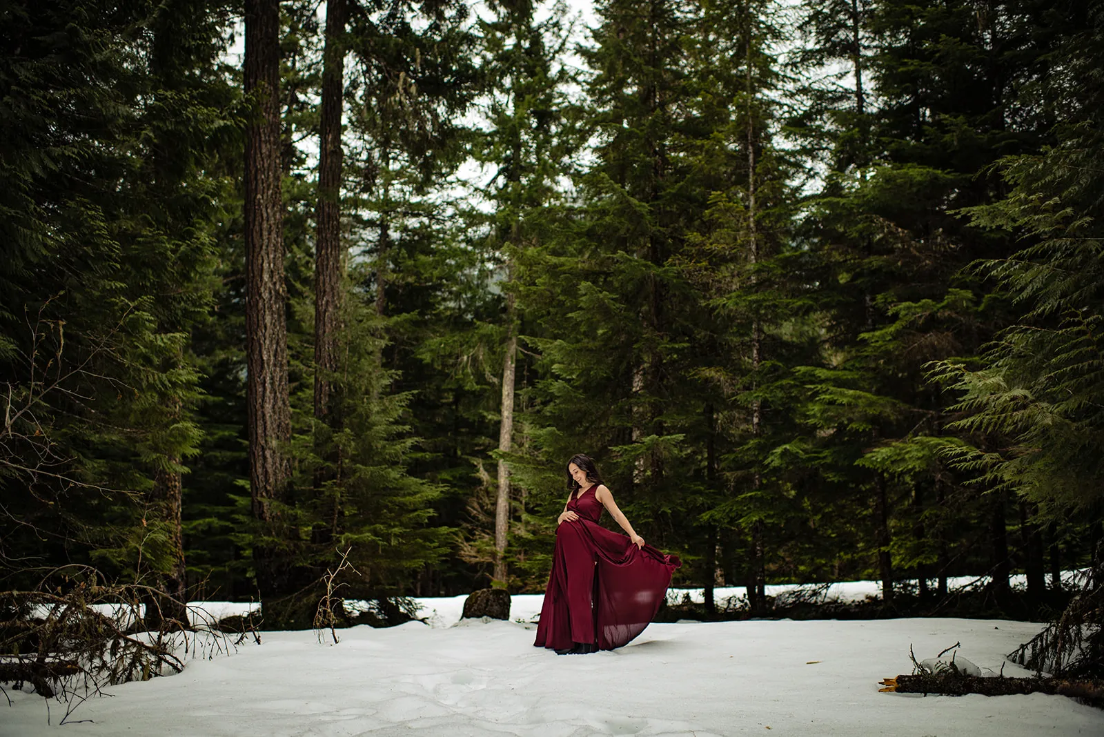 Pregnant woman in a flowing burgundy dress stands on snow in a dense green forest.