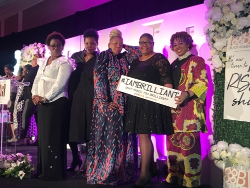 Group of five women posing on stage, one holding a sign reading #IAMBRILLIANT What makes you brilliant?
