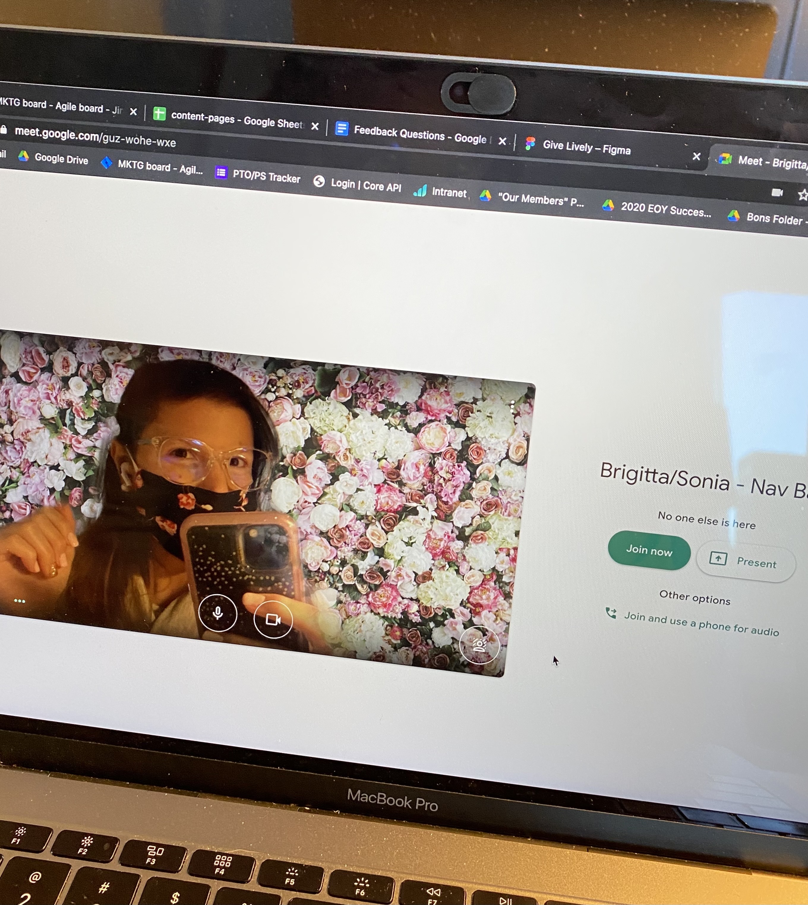 Screenshot of Sonia preparing to take part in a Google Meet. She's wearing a mask and glasses, and has a floral background.