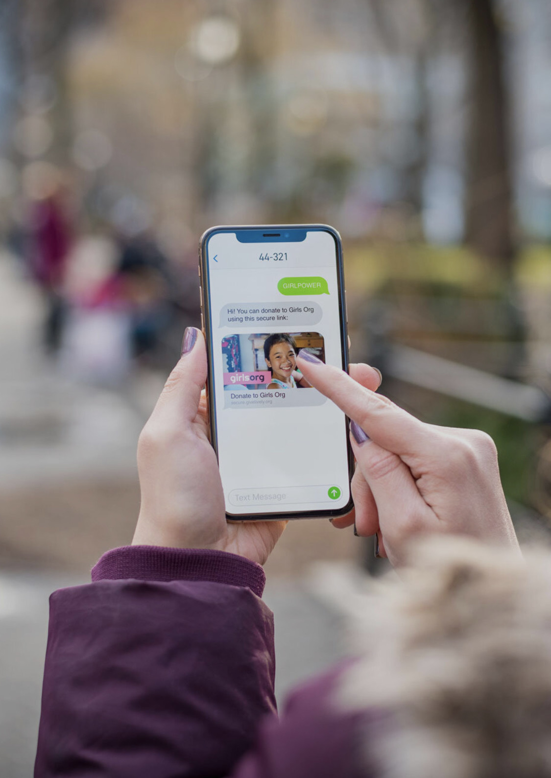 Two hands holding a mobile phone against a blurred outdoor background. The phone screen shows Text-to-Donate in action.