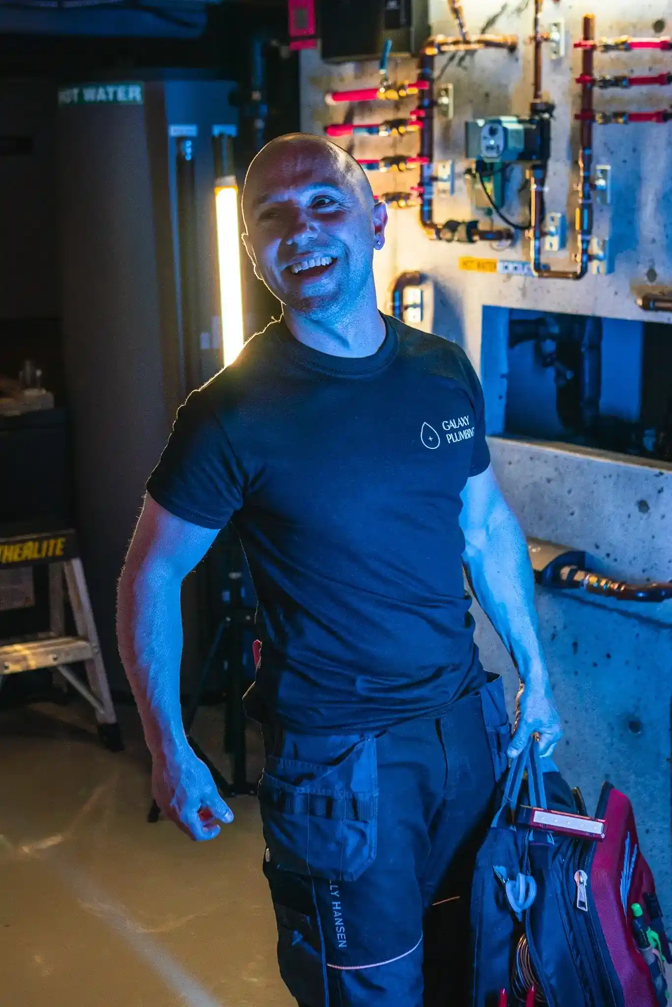 Smiling bald man in a black Galaxy Plumbing t-shirt standing in a utility room holding a toolbox bag, with pipes and equipment on the wall behind him.