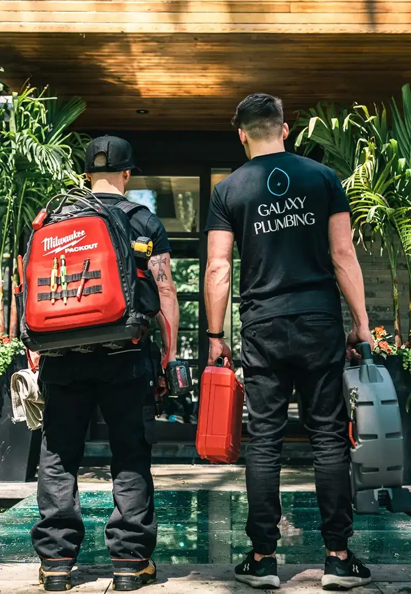 Two plumbers standing with their backs to the camera in front of a house entrance, one wearing a black cap and carrying a red Milwaukee Packout backpack with tools attached and the other wearing a black Galaxy Plumbing t-shirt holding two tool cases.