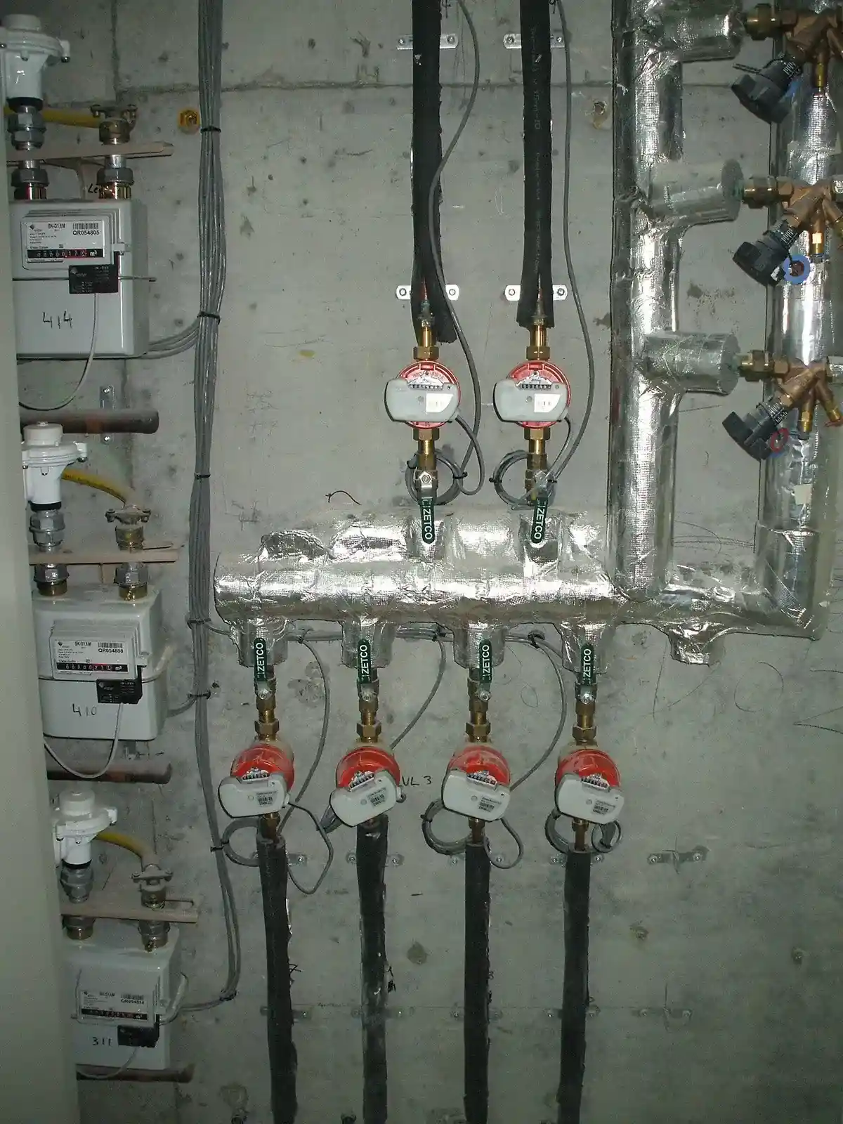 A wall-mounted plumbing setup with multiple water meters connected to insulated pipes and valves, against a concrete wall with visible wiring and utility labels.