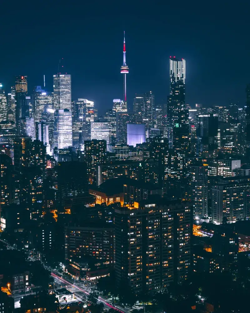 Nighttime cityscape with densely packed illuminated buildings and skyscrapers, featuring a tall tower with red and white lighting near the center.