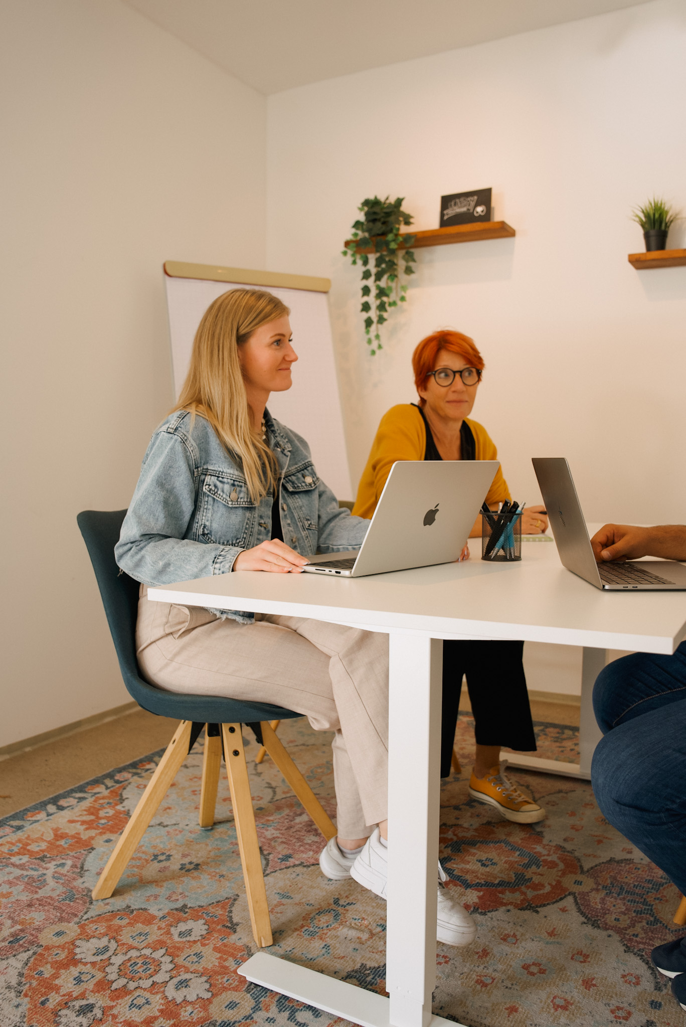 Zwei Frauen sitzen mit Laptops an einem weißen Tisch in einem hellen Besprechungsraum und hören aufmerksam zu. Am rechten Bildrand ist eine dritte Person angeschnitten, im Hintergrund stehen ein Flipchart und Wandregale mit Pflanzen.
