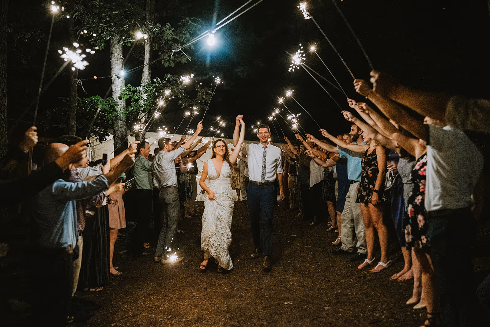 Bride and groom walking hand-in-hand through a pathway of guests holding sparklers at night.