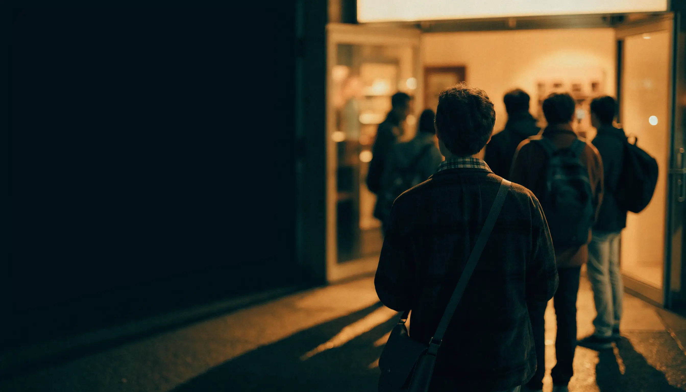 A short line of people waiting outside a shop door at night, seen from behind, representing customers who were already on their way
