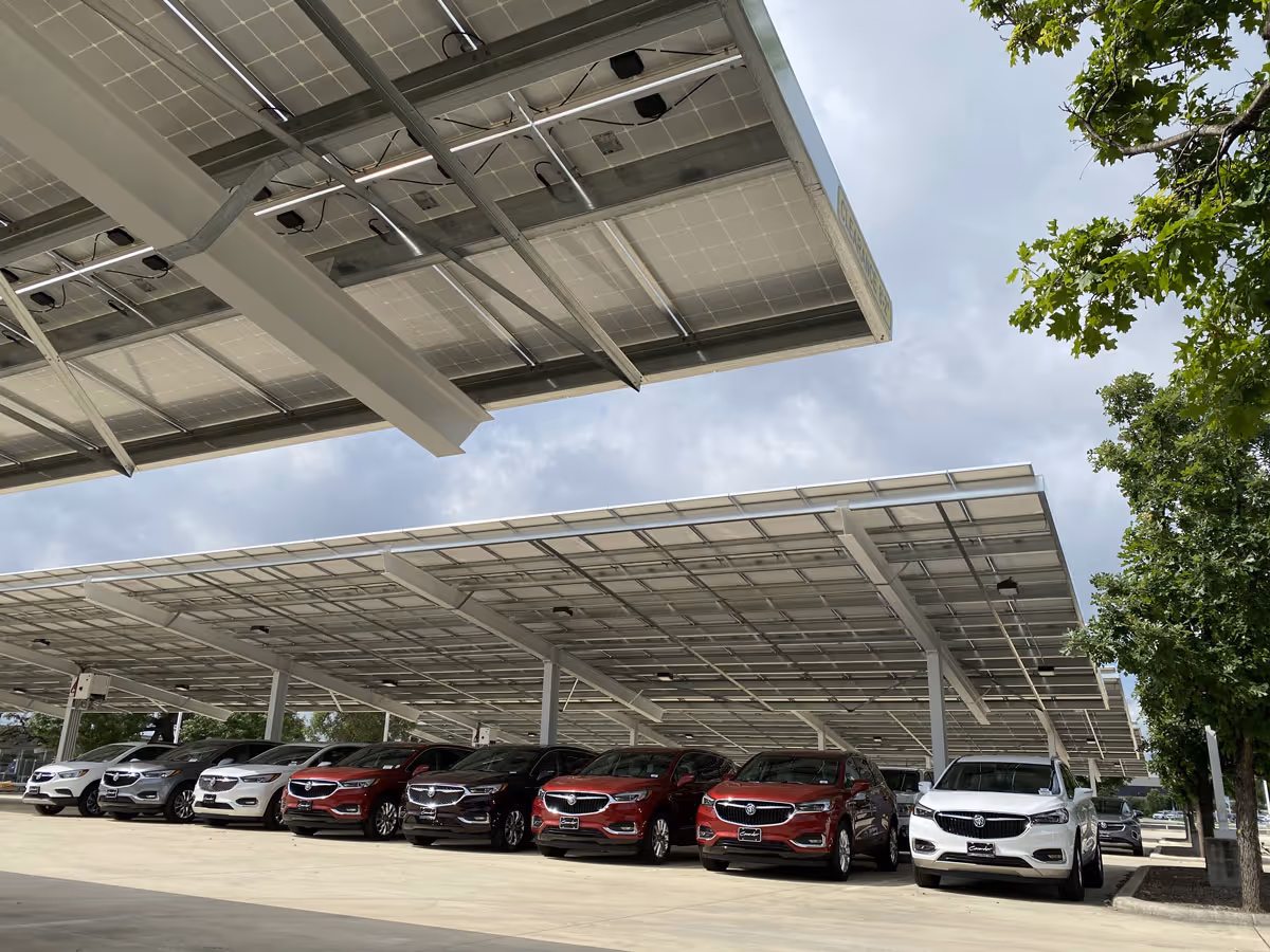 Row of parked Buick SUVs under a large solar panel carport with a cloudy sky and green trees nearby.