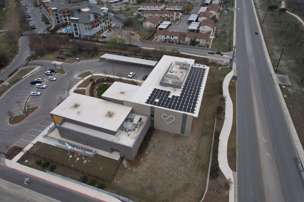 Aerial view of a modern building complex with solar panels on the roof and a heart-shaped design on one side, adjacent to a main road.