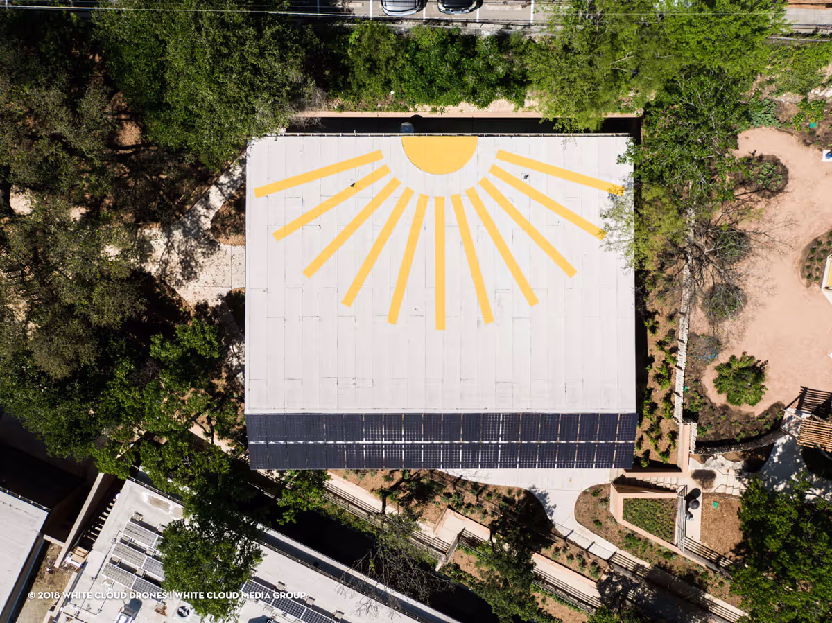 Aerial view of a building with a white roof featuring a yellow sun and rays design and solar panels on one side, surrounded by trees and pathways.