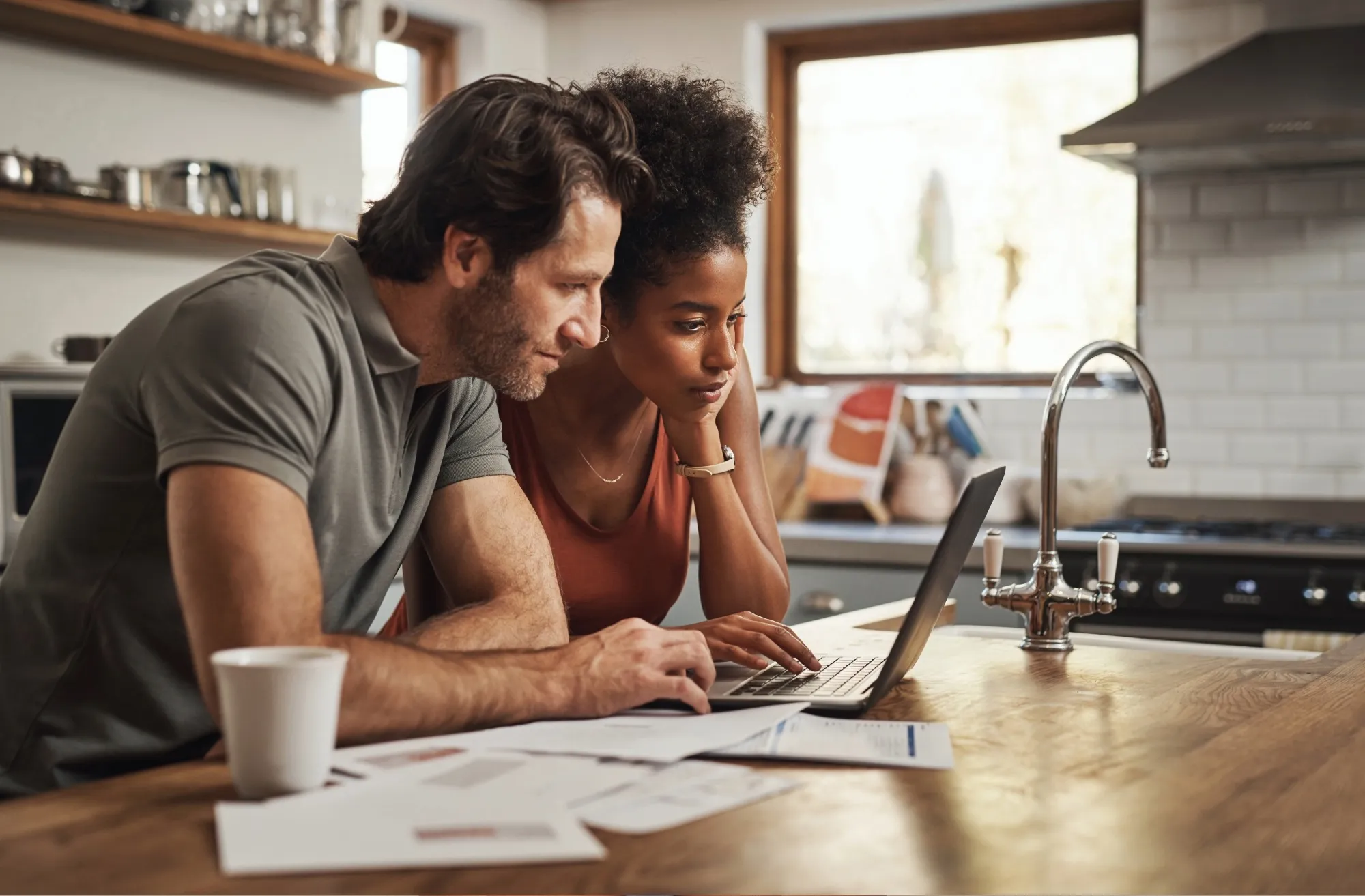 Couple working together on a laptop in a modern kitchen, surrounded by documents and a coffee cup.