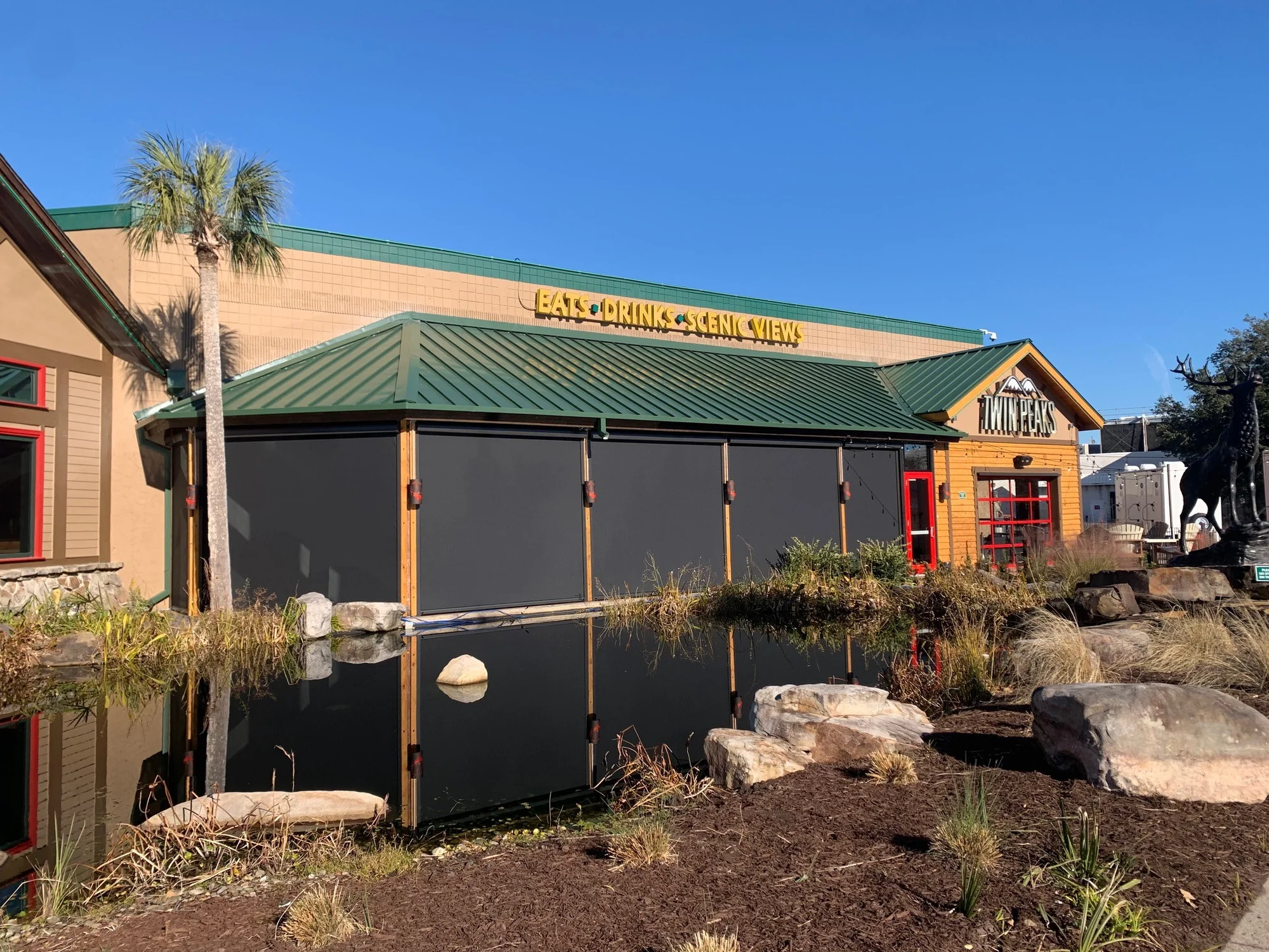 Exterior of Twin Peaks restaurant with a green roof and a pond reflecting the building and a nearby palm tree under clear blue sky.