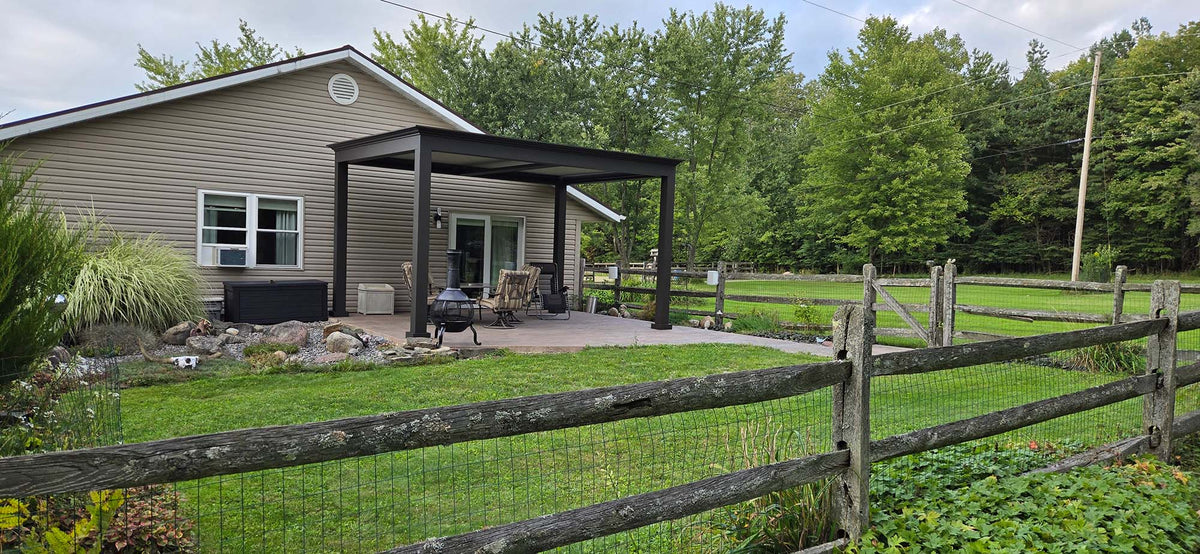 Backyard of a house with a covered patio, outdoor fireplace, lawn, and wooden fence under a cloudy sky.