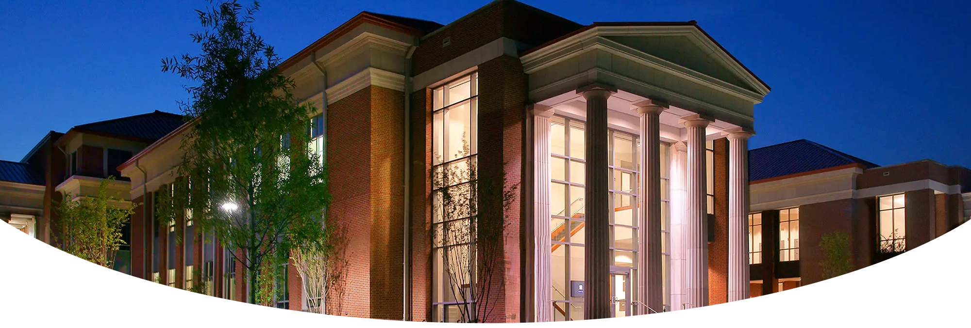 The University of Mississippi School of Law building at dusk, featuring large white columns, tall glass windows, and red-brick exterior walls illuminated by warm interior lighting.