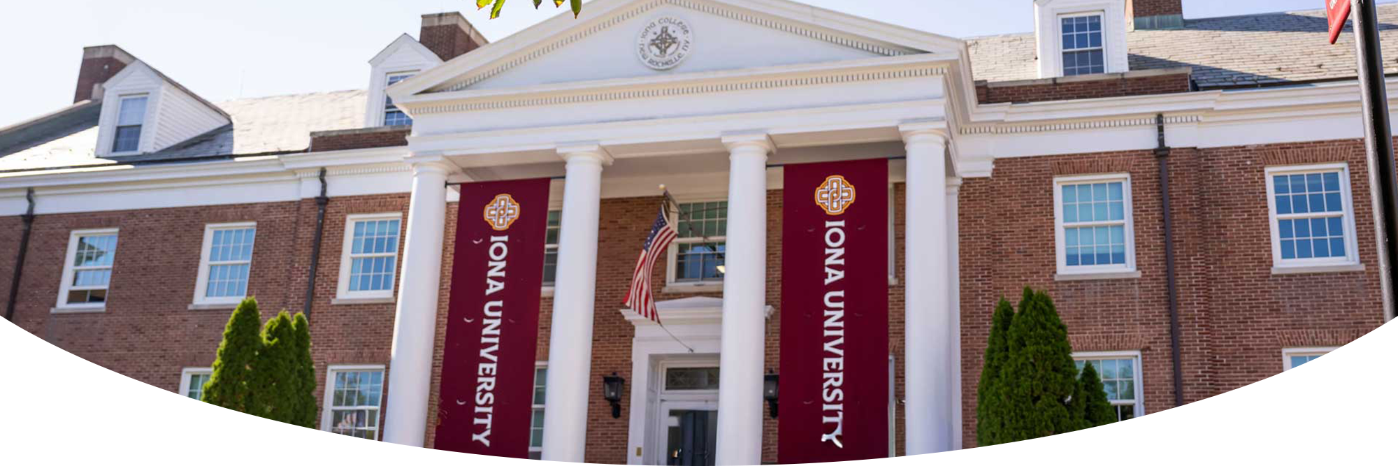 A bright, low-angle exterior photograph of a main building at Iona University on a sunny day. The red-brick structure features a grand white entrance portico with tall classical columns. Two large maroon vertical banners reading "IONA UNIVERSITY" hang prominently between the columns, flanking an American flag. Green tree leaves frame the top of the image, and wide stone steps lead up to the entrance.