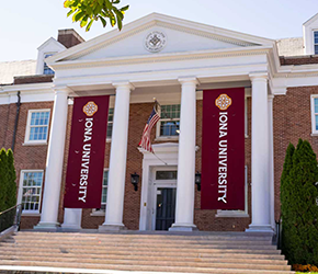 A bright, low-angle exterior photograph of a main building at Iona University on a sunny day. The red-brick structure features a grand white entrance portico with tall classical columns. Two large maroon vertical banners reading "IONA UNIVERSITY" hang prominently between the columns, flanking an American flag. Green tree leaves frame the top of the image, and wide stone steps lead up to the entrance.