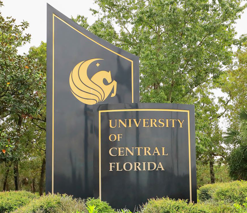 A black and gold University of Central Florida entrance sign featuring the Pegasus logo, surrounded by lush green trees.