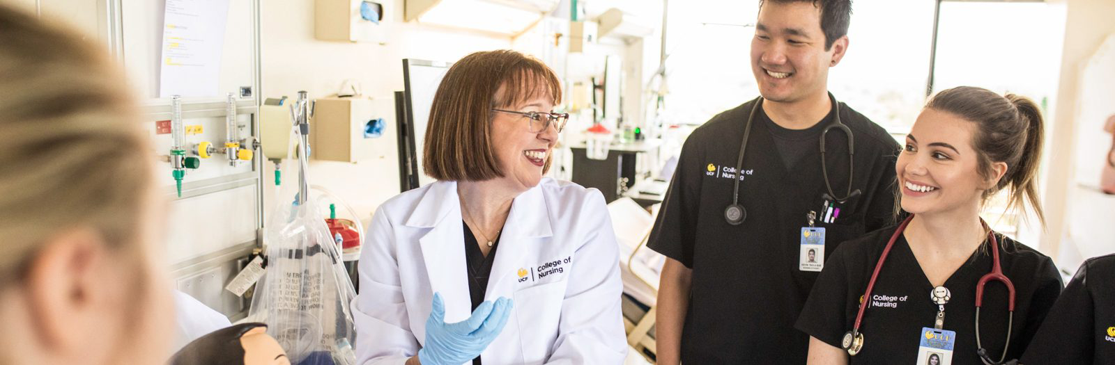 A smiling UCF College of Nursing instructor in a white lab coat teaching engaged nursing students in a clinical simulation lab.