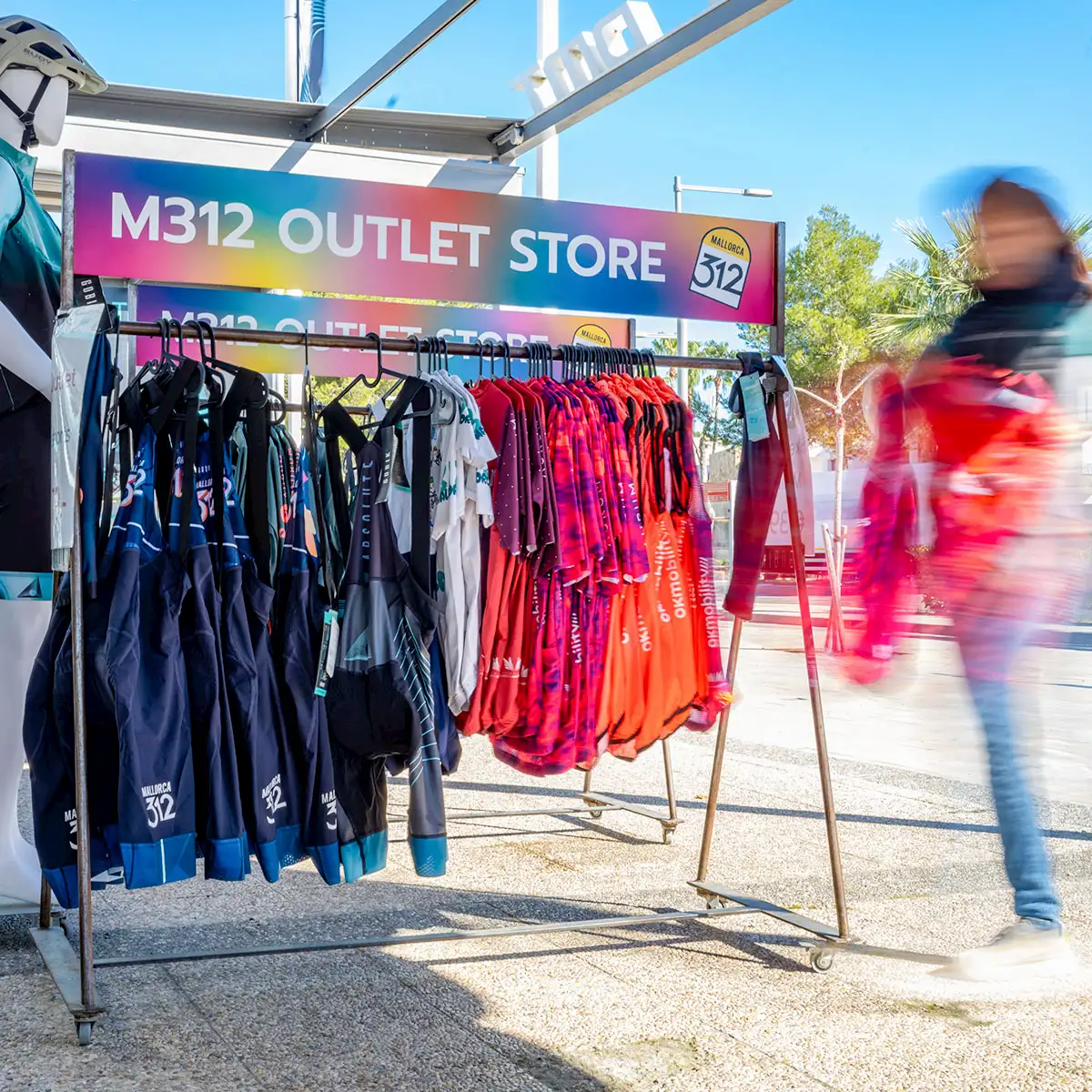 Kleiderständer mit farbigen Radtrikots und Sportbekleidung vor einem „M312 Outlet Store“, daneben eine vorbeigehende, verschwommene Person.