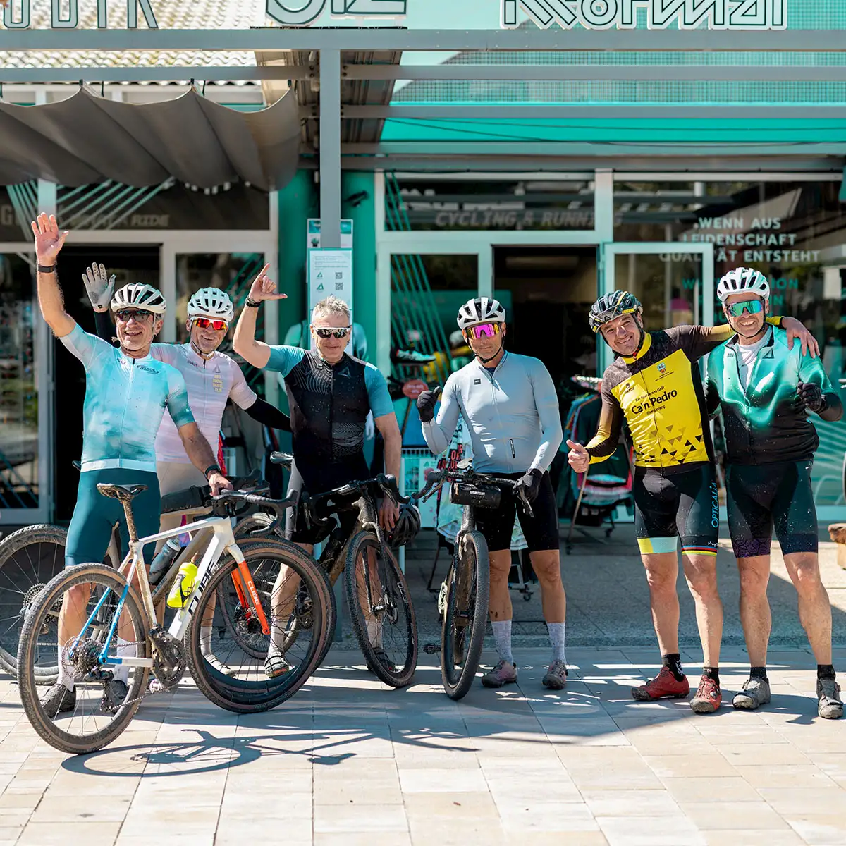 Gruppe von sechs Rennradfahrern in Sportbekleidung steht vor einem Fahrradgeschäft, hält ihre Bikes und posiert gut gelaunt nach einer Ausfahrt.