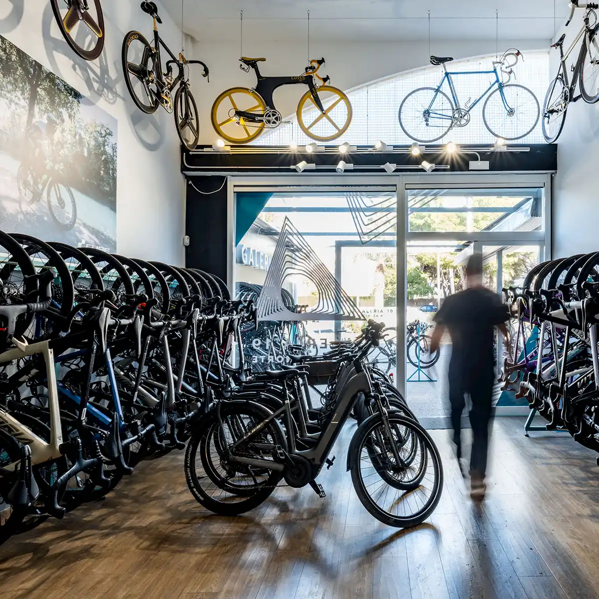 Innenansicht eines Fahrradgeschäfts mit Reihen von E-Bikes und Rennrädern, während eine Person unscharf durch den Laden in Richtung Ausgang geht.