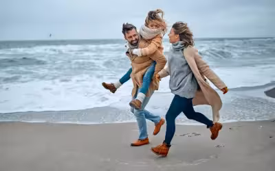 photo of a happy family running on the beachfront wearing warm clothes the child is being carried on the fathers back
