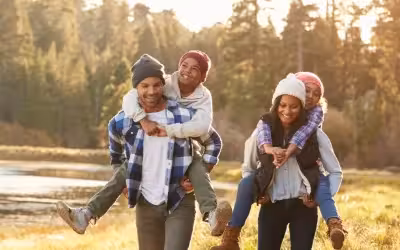 Family out walking by a lake and trees wearing winter woolly hats, daughter is being carried on Mums back and son on Dads back. Life Insurance Payouts: How Long Do They Take in the UK?