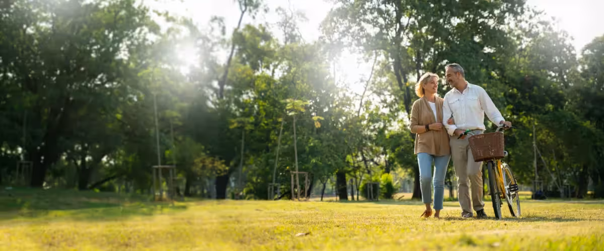 Older couple walking through the park with trees and sunshine in background, man is pushing a bicycle alongside. How Does Life Insurance With Terminal Illness Cover Work?