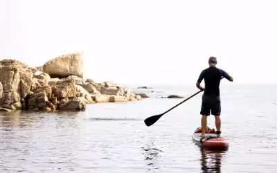 older man kneeling on a paddleboard in a calm sea with rocky coastline