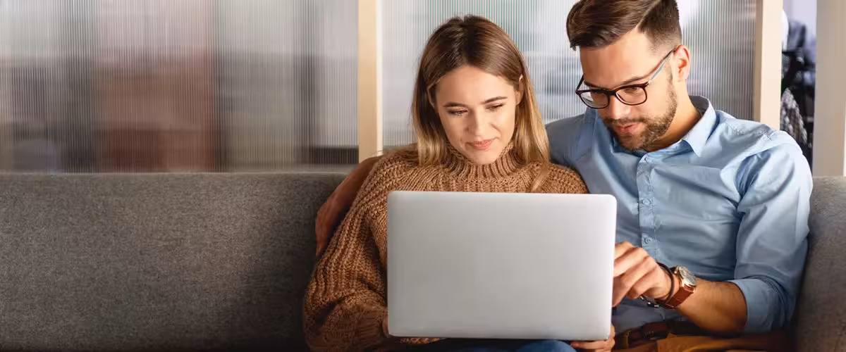 Young couple sitting on their sofa looking at a laptop reading about why health insurance is so complicated in the UK