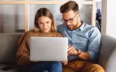 Young couple sitting on their sofa looking at a laptop reading about why health insurance is so complicated in the UK