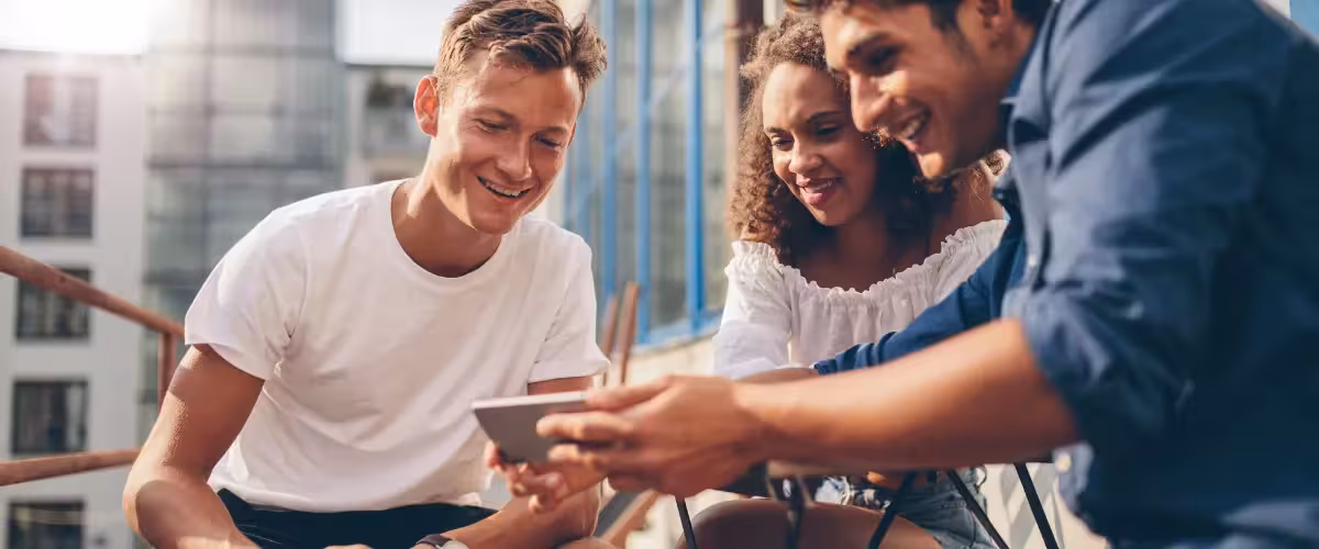 Group of young adults gathered around phone smiling. What Are the Benefits Of Getting Health Insurance While You're Young?