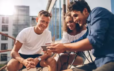 Group of young adults gathered around phone smiling. What Are the Benefits Of Getting Health Insurance While You're Young?