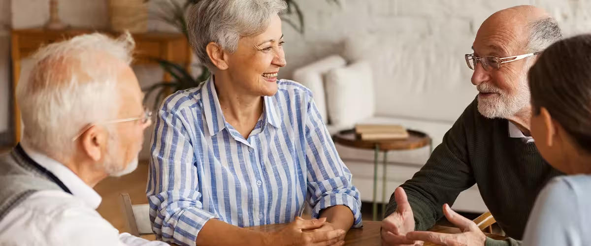 Group of four cheerful senior friends, two men and two women, sitting at table and enjoying talk after playing cards in assisted living home. 14 Ways Over 65s Can Reduce The Cost of Private Health Insurance