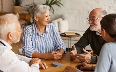 Group of four cheerful senior friends, two men and two women, sitting at table and enjoying talk after playing cards in assisted living home. 14 Ways Over 65s Can Reduce The Cost of Private Health Insurance