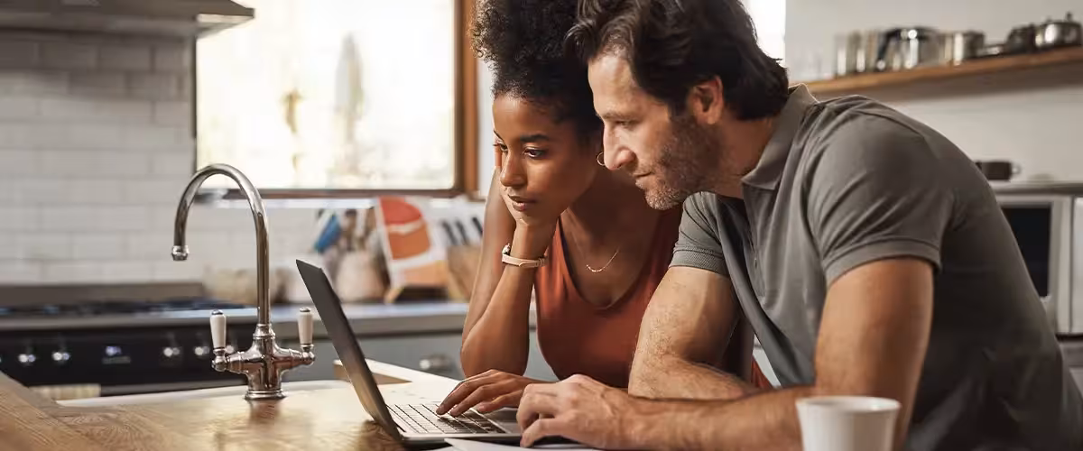 Couple looking at their laptop in the kitchen with documents on the side 