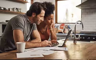 Couple looking at their laptop in the kitchen with documents on the side 