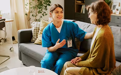 A healthcare professional in scrubs speaks reassuringly with a woman on a sofa at home, illustrating access to private healthcare services in the UK.