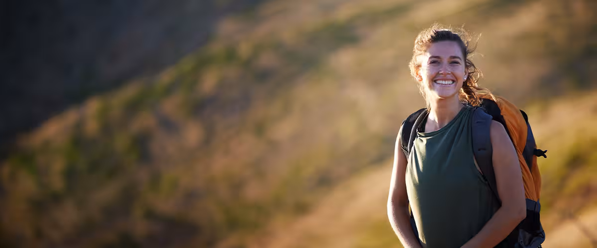 Smiling woman with a backpack hiking on a sunlit grassy hillside