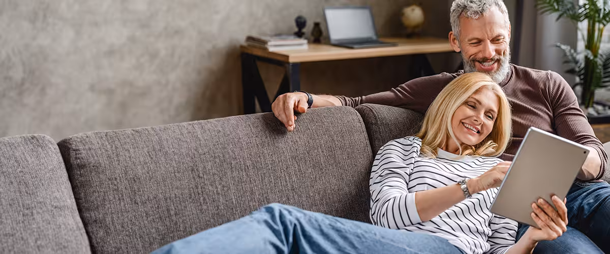 A relaxed older couple sitting on a sofa at home, smiling while using a tablet together, representing reviewing and managing health insurance options for over 50s