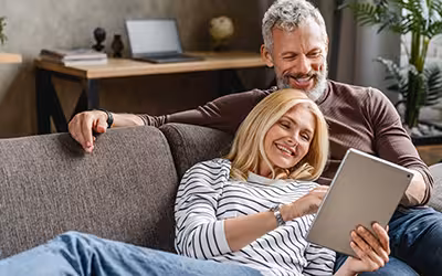 A relaxed older couple sitting on a sofa at home, smiling while using a tablet together, representing reviewing and managing health insurance options for over 50s