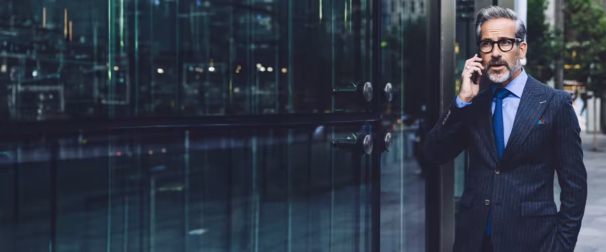 A well-dressed professional man in a suit talking on his phone outside a modern office building, representing premium private health insurance for high-net-worth individuals and global professionals