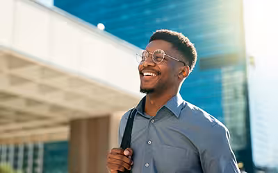 Smiling man with glasses and a shoulder bag stands outside a modern office building