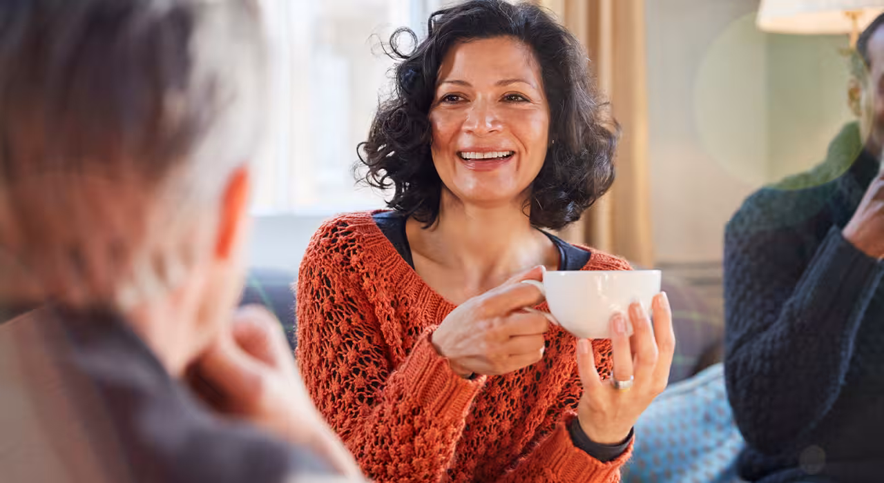 Smiling woman holding a cup of tea while chatting with others at home, reflecting supportive conversations around menopause and wellbeing.