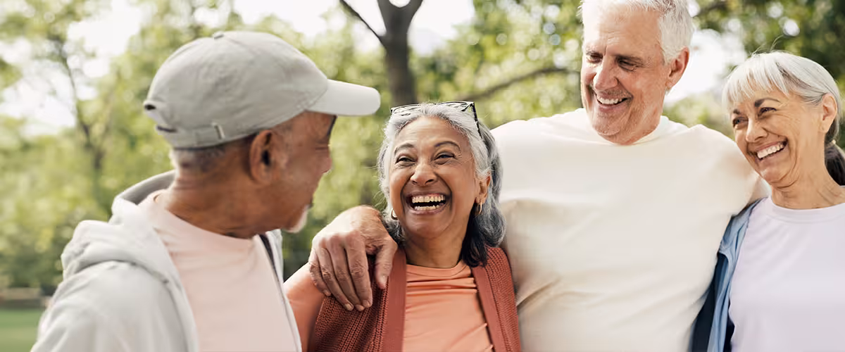 Four smiling older adults stand close together outdoors, laughing and embracing in a sunlit park.