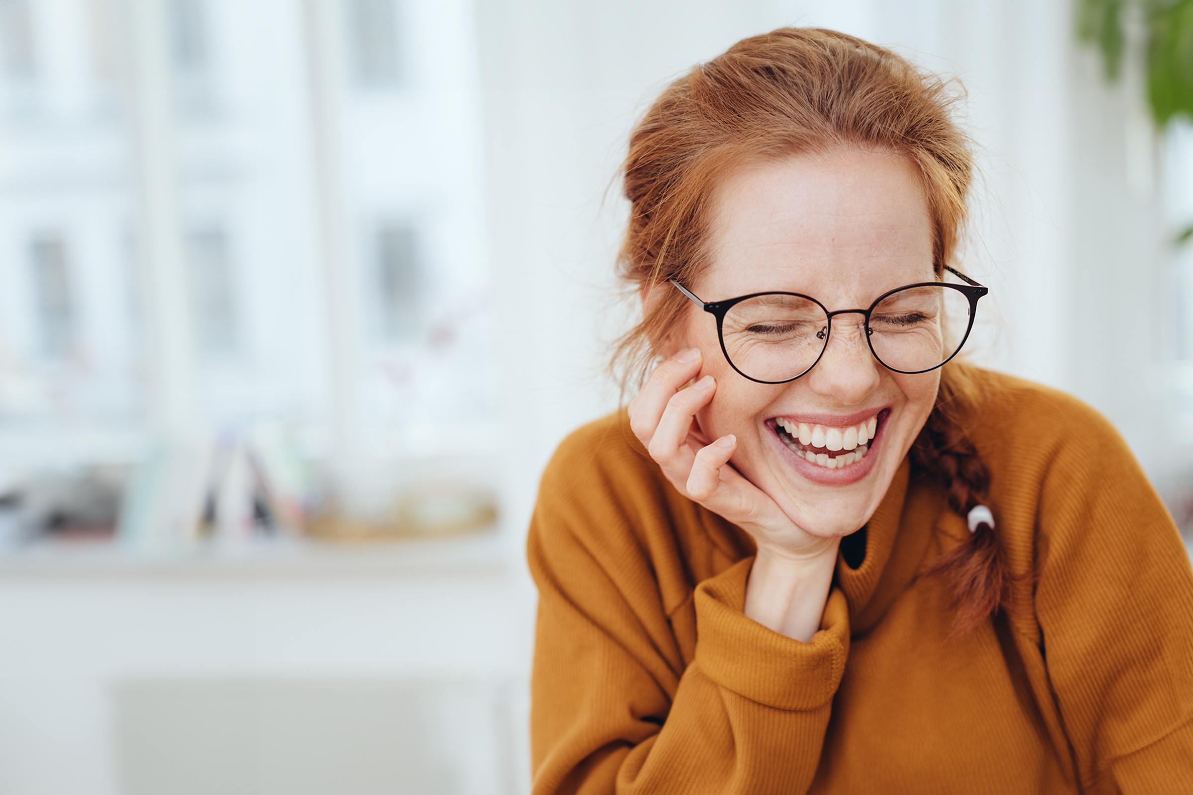 Woman in glasses laughing with her hand resting against her cheek in a bright room